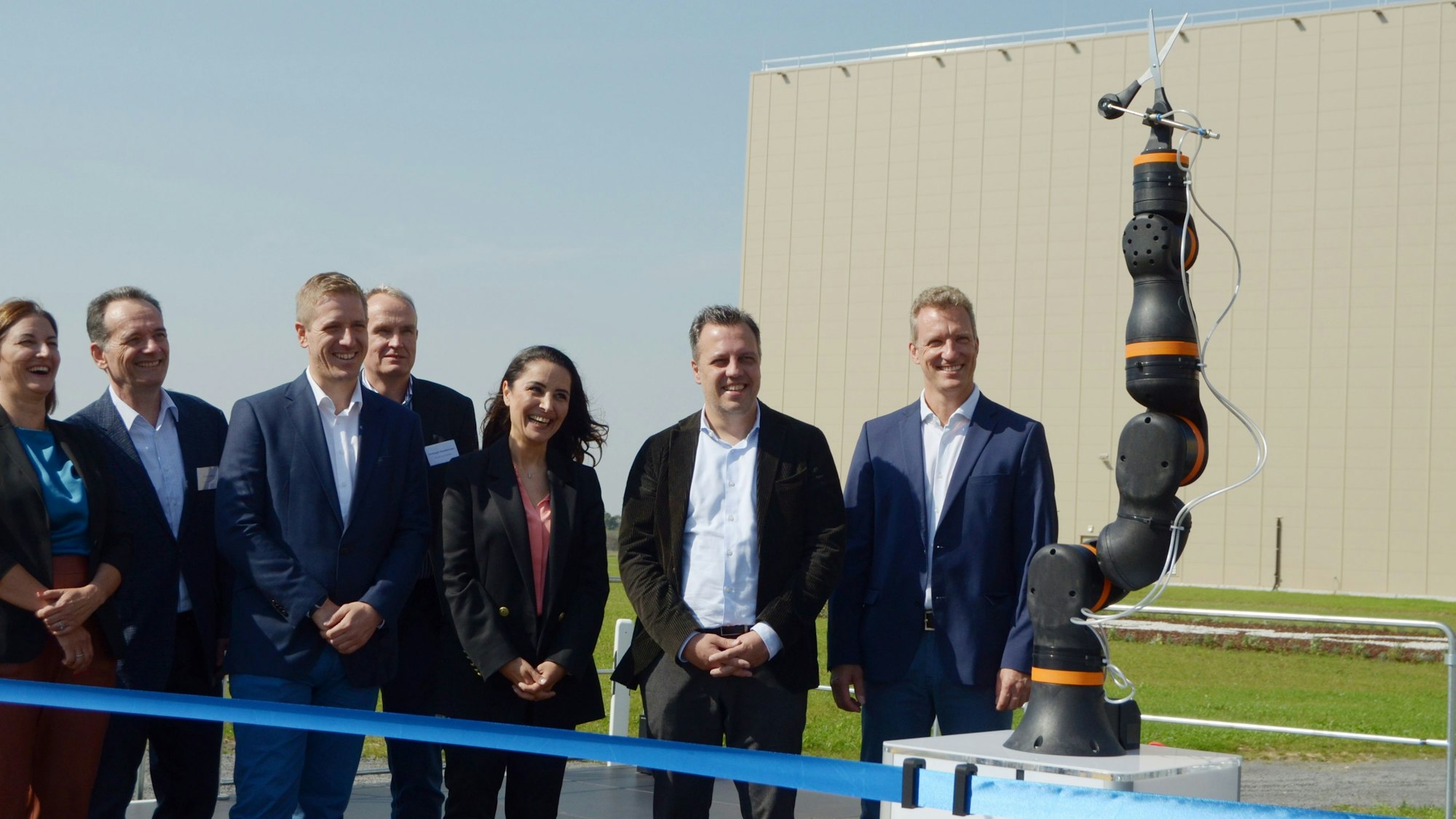 Vertreter des Unternehmens Procter & Gamble und Gäste stehen hinter einem blauen Band. Sie beobachten, wie ein Roboter eine Schere in Position bringt, die das Band durchtrennen soll.