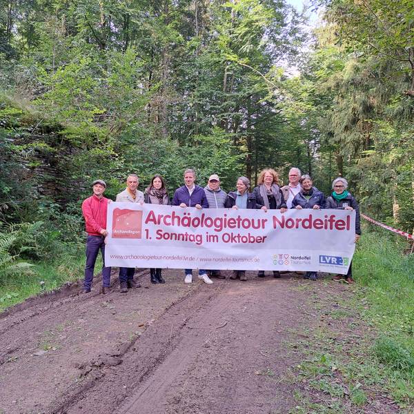 Eine Gruppe Menschen steht im Wald und hält ein Transparent, das für die Archäologietage wirbt.
