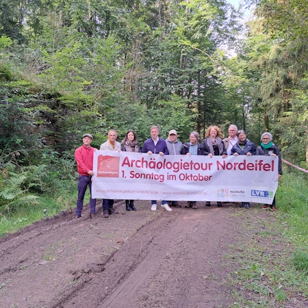 Eine Gruppe Menschen steht im Wald und hält ein Transparent, das für die Archäologietage wirbt.