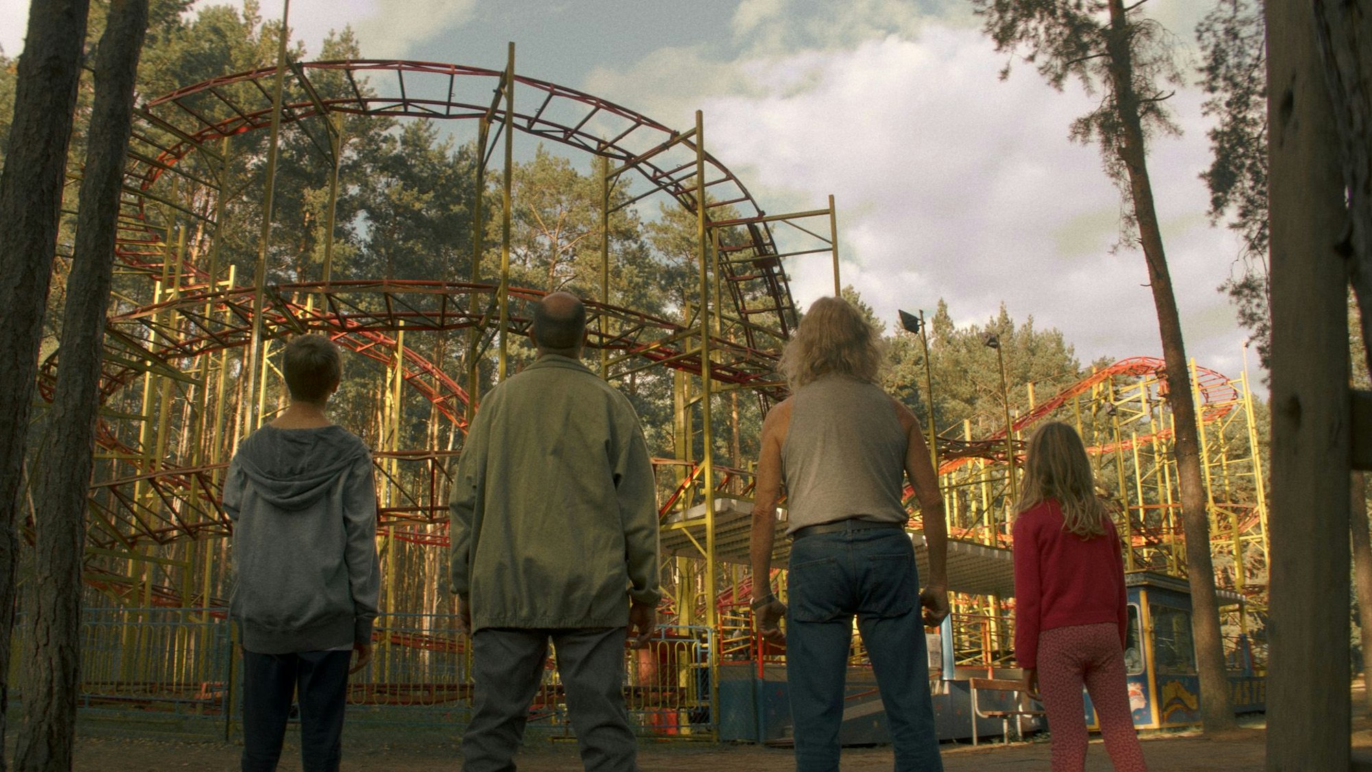Zwei Männer und zwei Kinder stehen vor einer maroden Achterbahn.