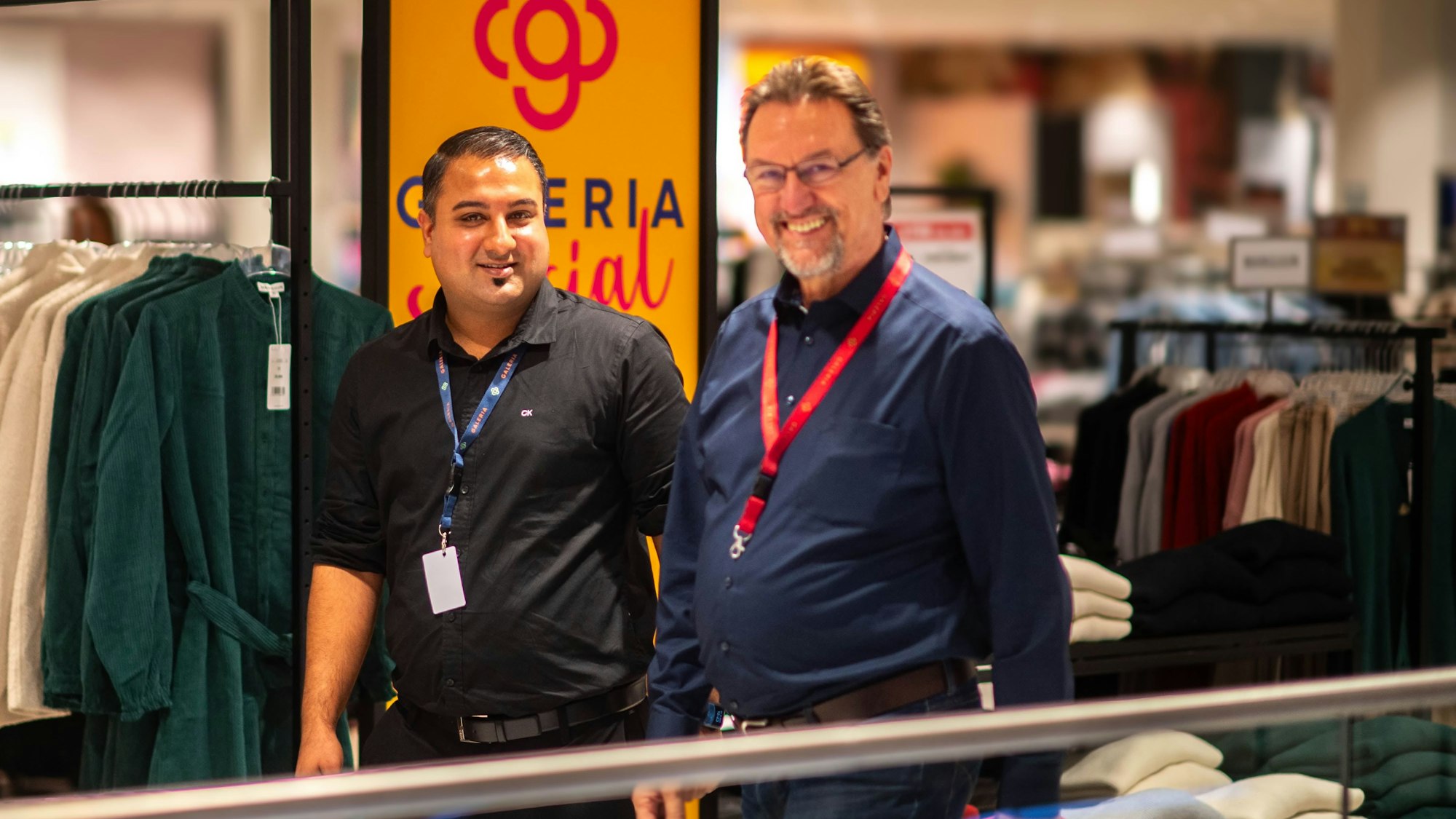Zwei Männer gehen durch ein Bekleidungsgeschäft und schauen fröhlich in die Kamera.