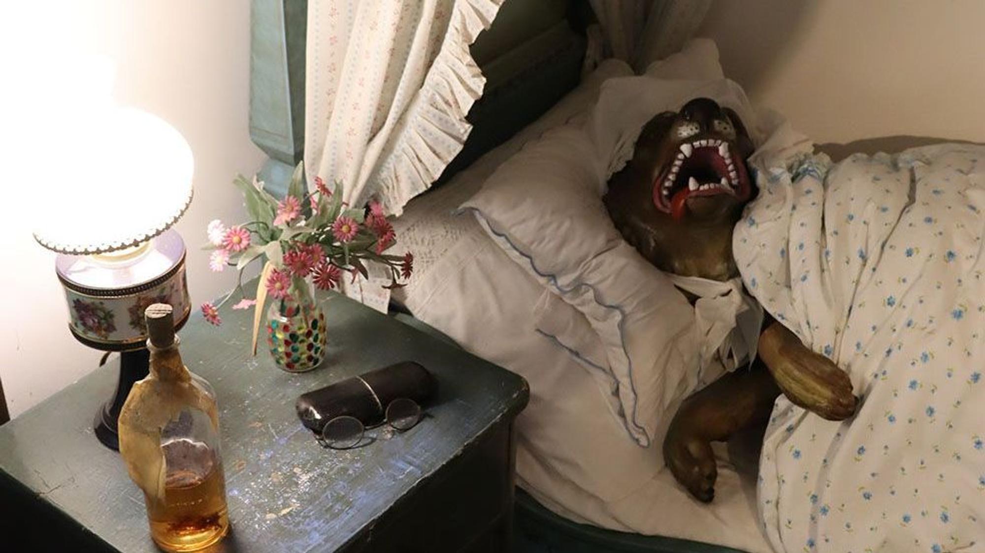 Eine Szene im Märchenwald: Der Wolf liegt als Großmutter verkleidet im Bett. Er sperrt das Maul auf und zeigt seine Zähne.