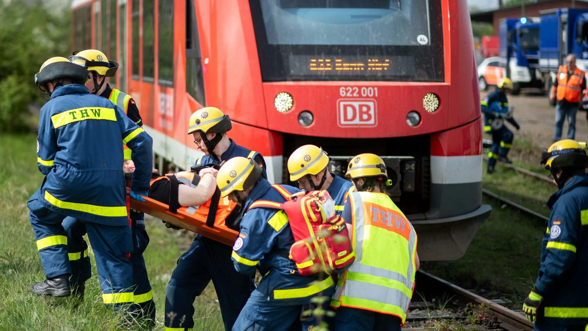 Helfer des THW tragen bei einer Übung einen Verletztendarsteller auf einer Trage weg. Im Hintergrund ist ein roter Zug zu sehen.