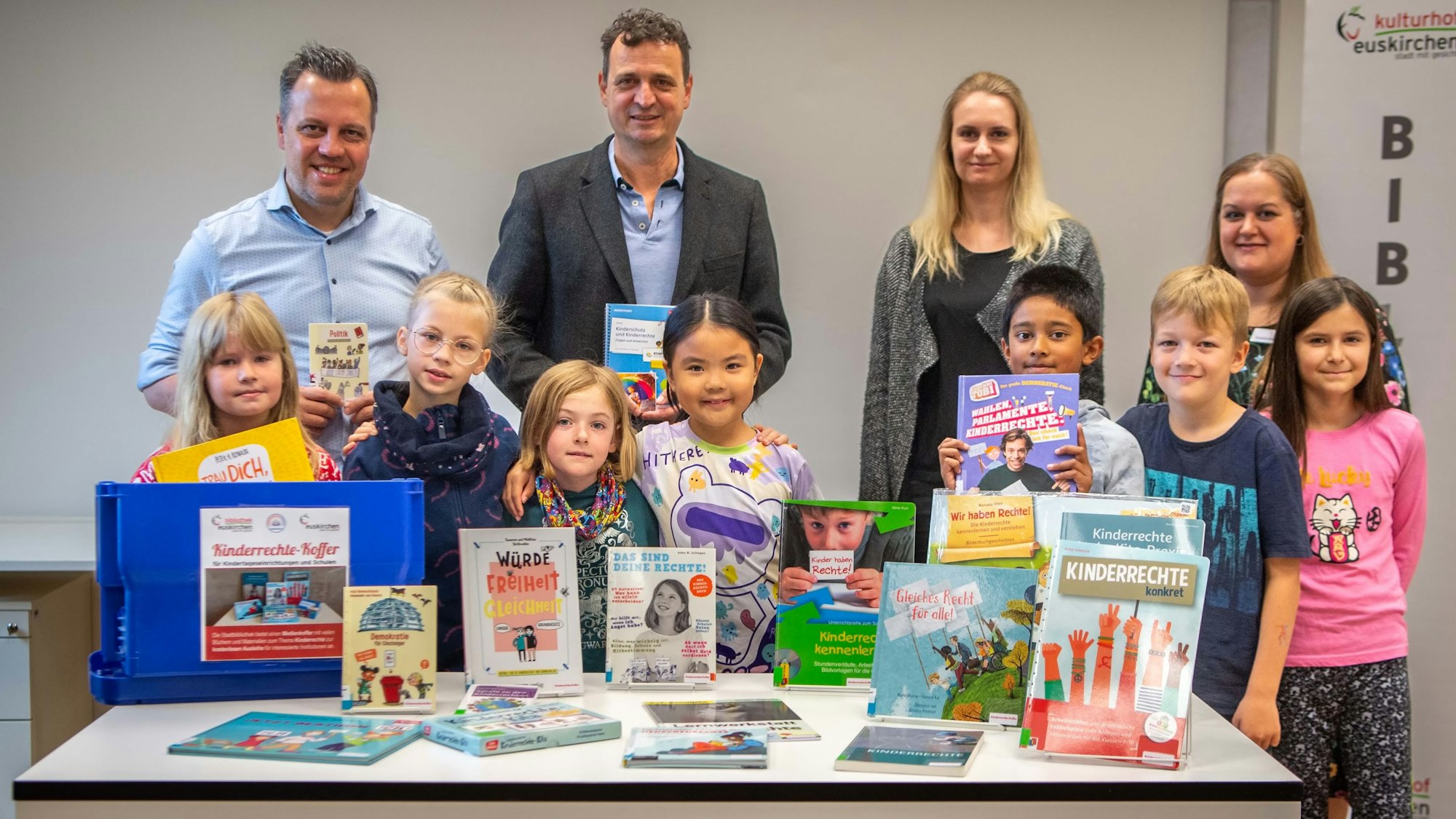 Das Bild zeigt zahlreiche Bücher, die auf einem Tisch stehen und liegen. Dahinter stehen Kinder und Erwachsene.
