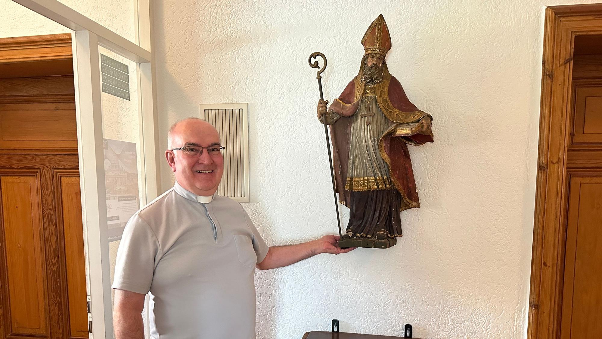 Lambert Schäfer steht neben einer Heiligenfigur, er trägt ein hellgraues Hemd mit römischem Kragen.
