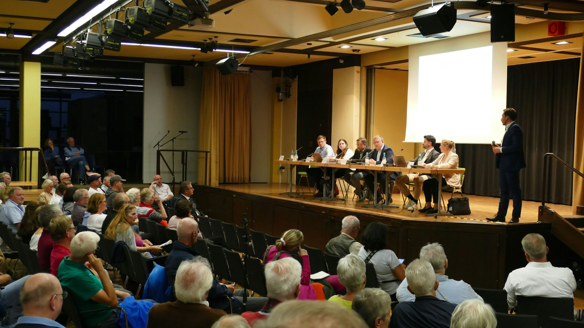Besucher einer Informationsveranstaltung sitzen in einer Schulaula. Auf der Bühne sitzen Männer und Frauen an einem Podium.