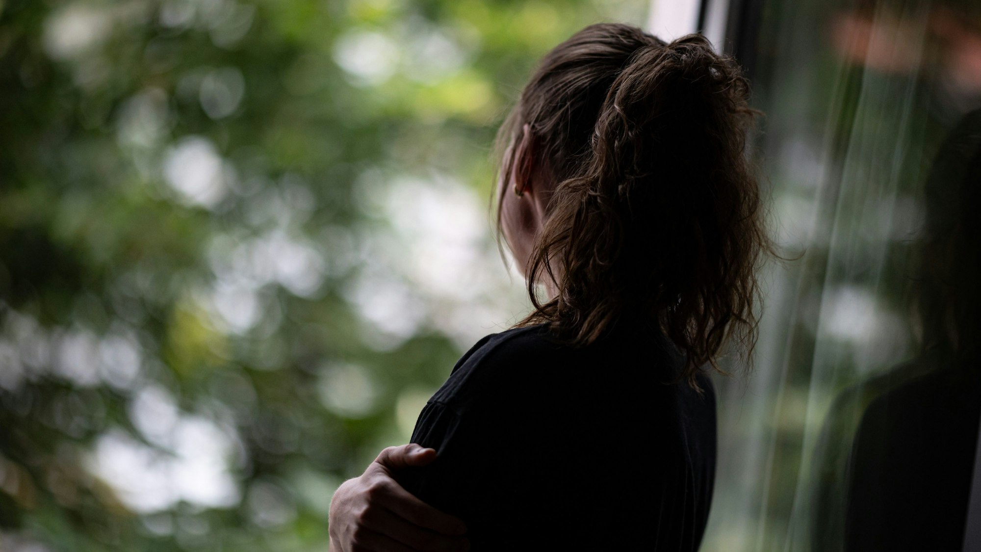 Eine junge Frau steht in einer Wohnung und schaut durch ein Fenster nach draußen.