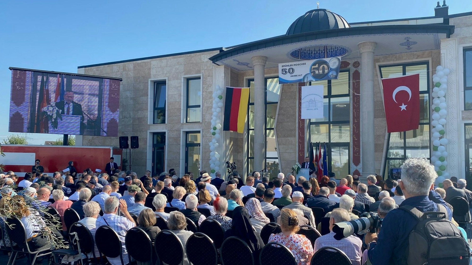 Zu sehen sind vollbesetzte Stuhlreihen vor der neuen Moschee.