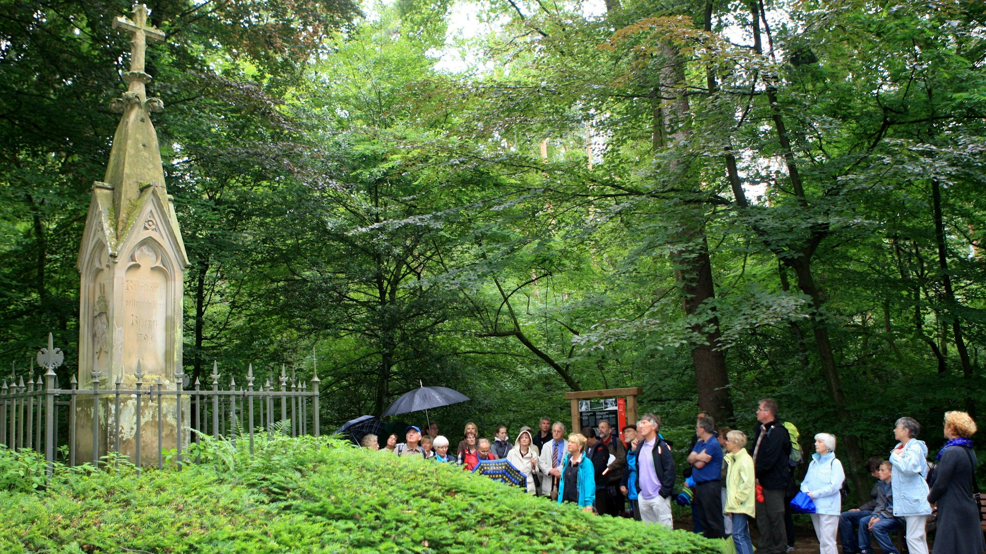 Eine Wandergruppe steht vor einem Denkmal mitten im Wald in der Mitte von Bergisch Gladbach.