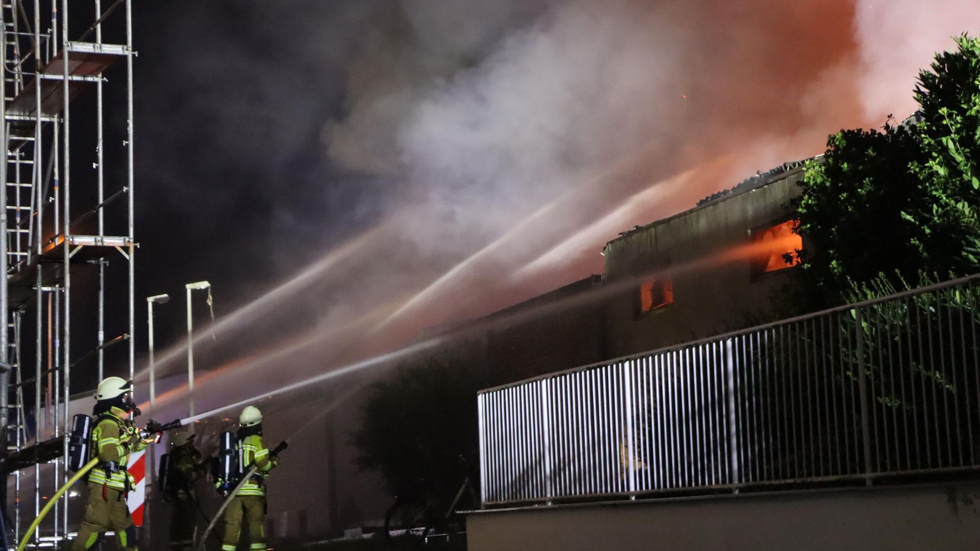 Das Bild zeigt mehrere Feuerwehrleute mit Löschschläuchen, die versuchen, die Flammen in einem Gebäude zu löschen.