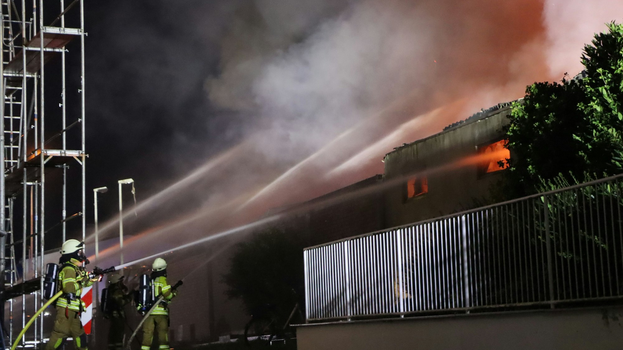 Das Bild zeigt mehrere Feuerwehrleute mit Löschschläuchen, die versuchen, die Flammen in einem Gebäude zu löschen.