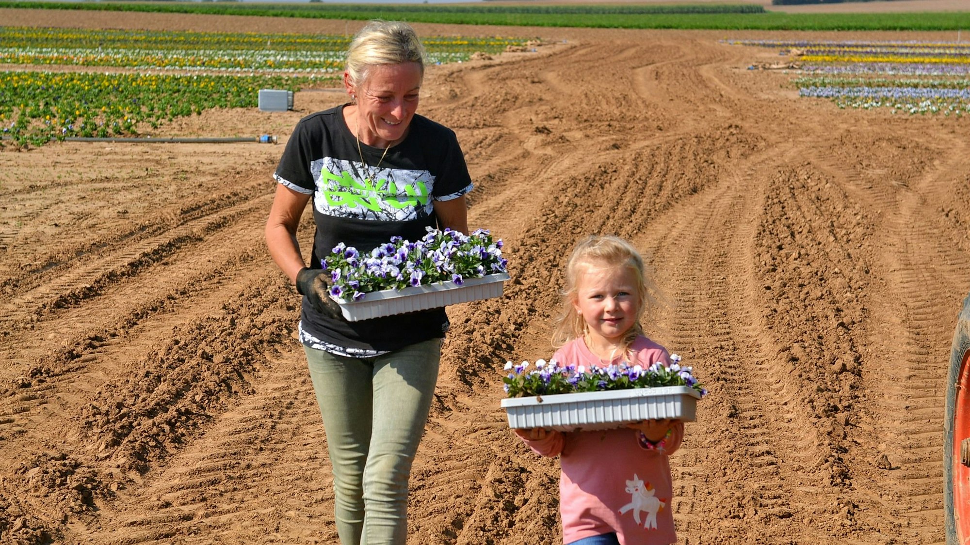 Hilfsarbeiterin Irena und Landwirtstochter Romy tragen eine Tablett mit Blumen.