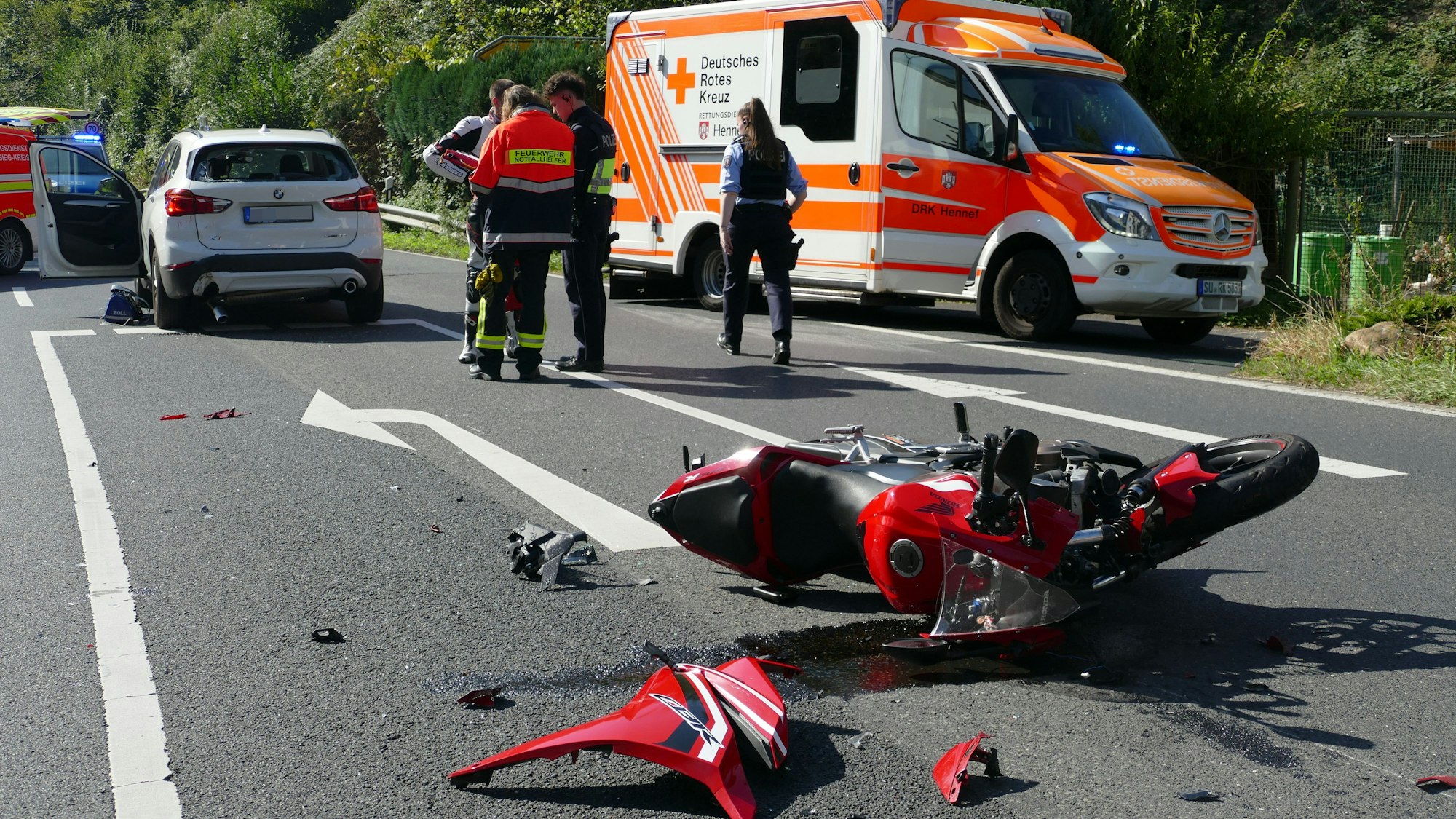 Ein rotes Motorrad liegt auf der Seite auf der Fahrbahn, Teile liegen daneben. Ein weißes Auto und ein Rettungswagen stehen dahinter.