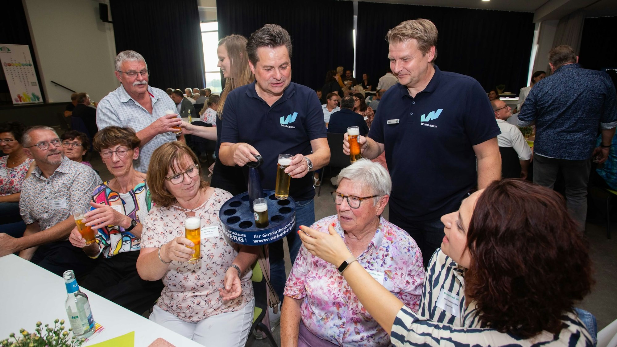 Die drei genannten Personen stehen mit einem Kölschkranz hinter sitzenden Damen.