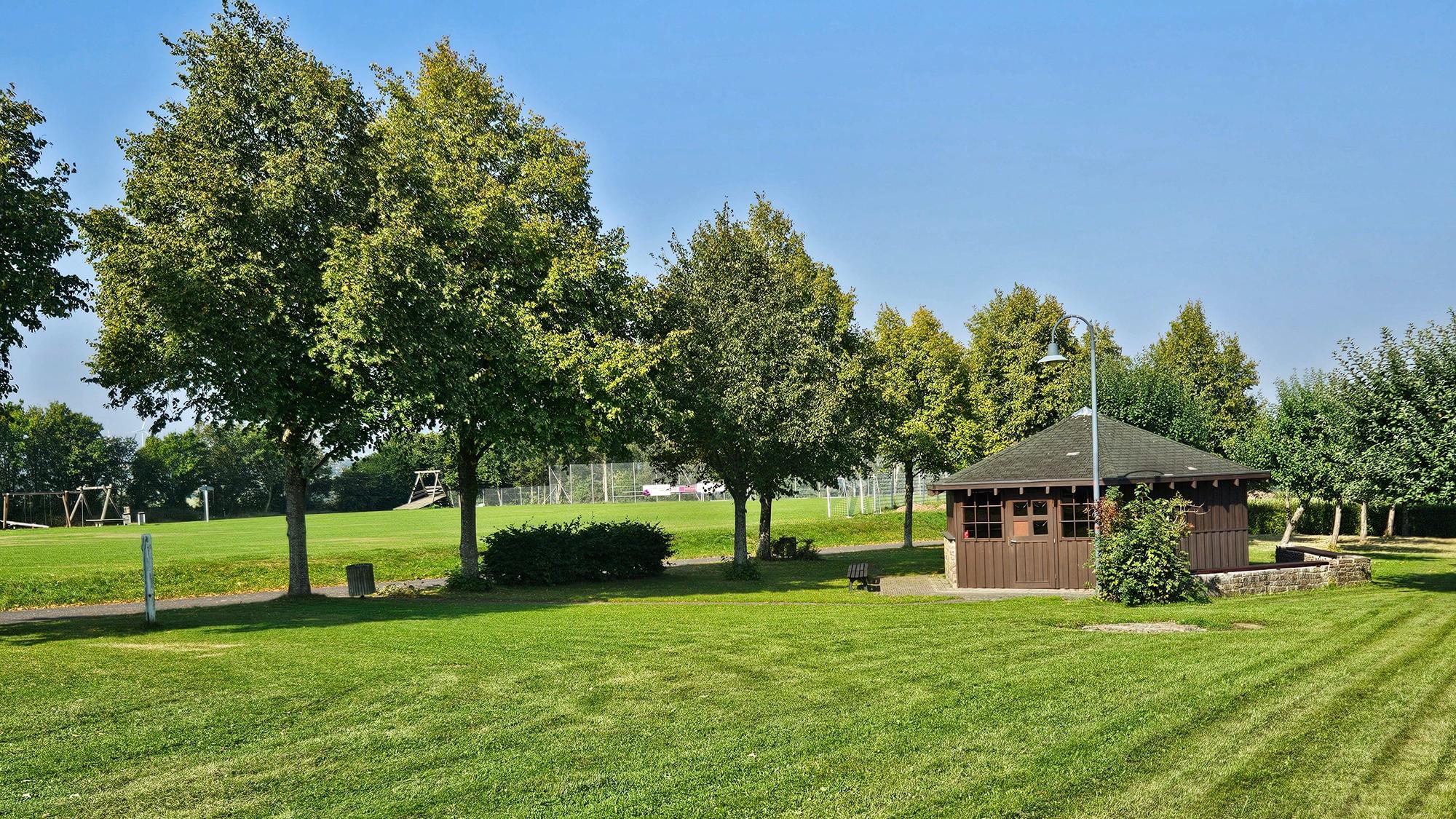 Blick auf die Wiese mit der Grillhütte und dem Sportplatz im Hintergrund.