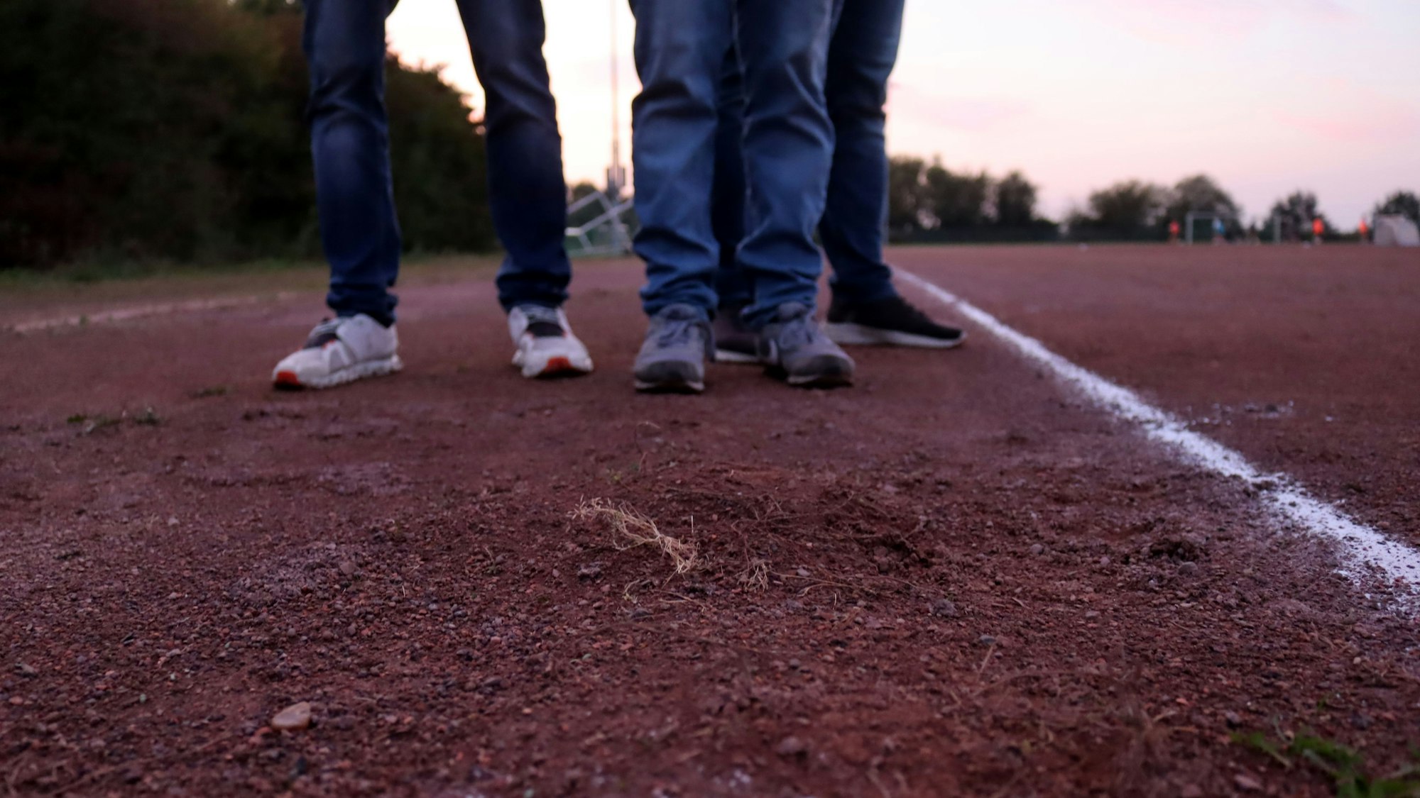 Am Rande des Tennenplatzes sind Unebenheiten zu erkennen. Im Hintergrund sind man die Beine von drei Personen.