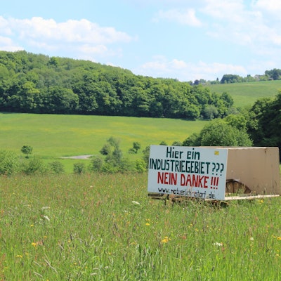 Auf einer großen Wiese mit einem bewaldeten Hügel im Hintergrund steht ein Protestplakat mit der Aufschrift „Hier ein Industriegebiet? Nein danke!“.