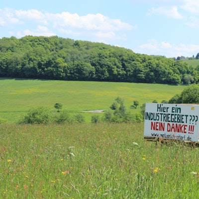 Eine Holztafel mit der Aufschrift Hier ein Industriegebiet? Nein danke! steht in einer hügeligen Wiesenlandschaft mit Bäumen im Hintergrund.