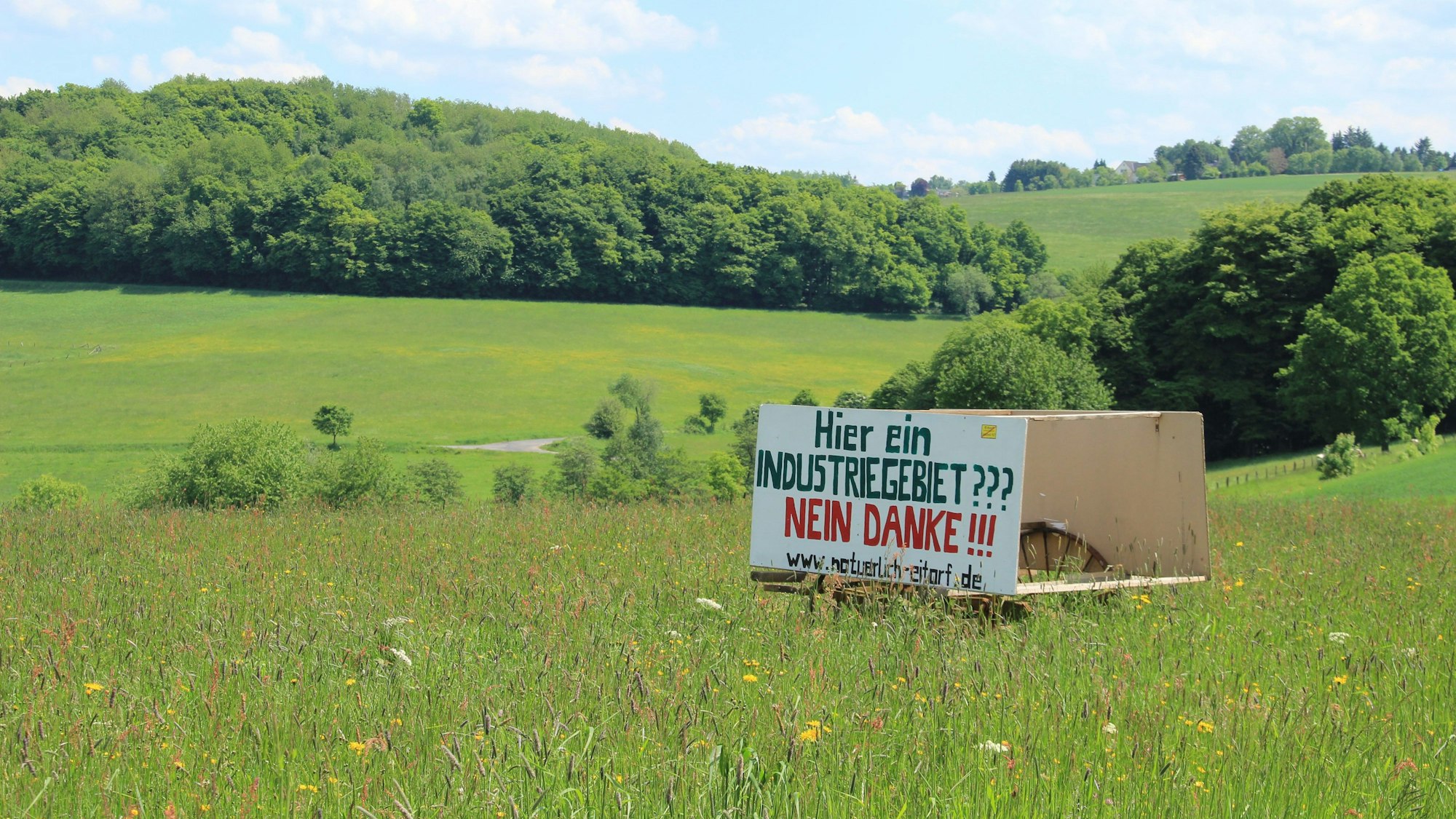 Eine Holztafel mit der Aufschrift Hier ein Industriegebiet? Nein danke! steht in einer hügeligen Wiesenlandschaft mit Bäumen im Hintergrund.