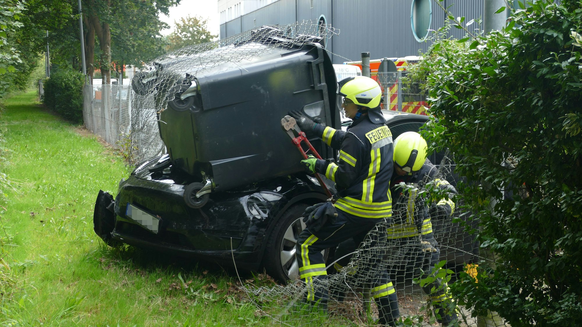 Zwei Feuerwehrleute stehen mit einer großen Zange neben einem schwarzen Tesla, auf dessen Motorhaube ein Müllcontainer liegt. Das Fahrzeug hat einen Maschendrahtzaun durchbrochen.