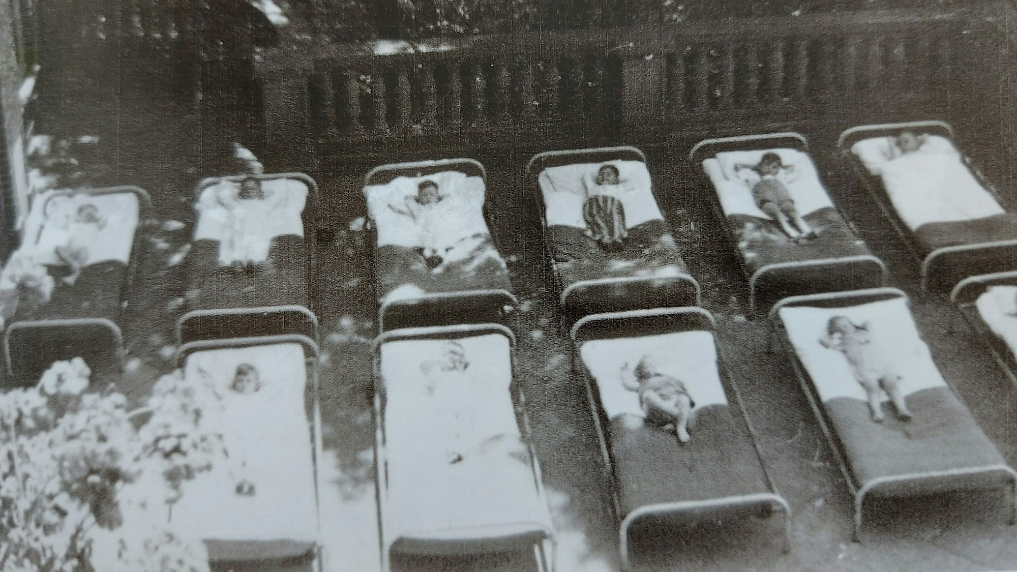 10 Kinder liegen in sorgsam aufgereihten Krankenbetten auf einer Terrasse.