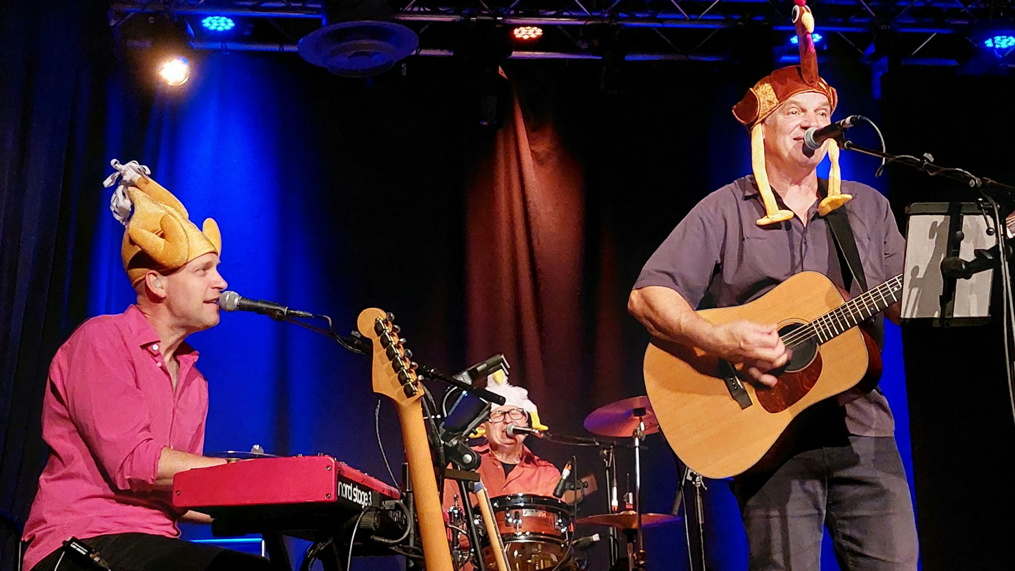 Drei Musiker mit lustigen Hühner-Kappen auf dem Kopf spielen Gitarre, Keyboard und Schlagzeug und singen.