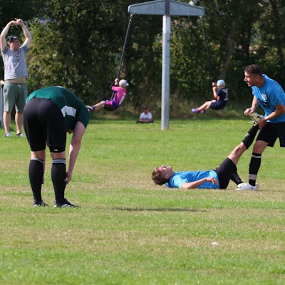 Ein Fußballer liegt mit einem Krampf auf dem Sportplatz, ein Mitspieler hilft ihm. Ein Gegenspieler beugt sich nach vorne. Im Hintergrund gibt der Trainer das Zeichen zum Auswechseln.