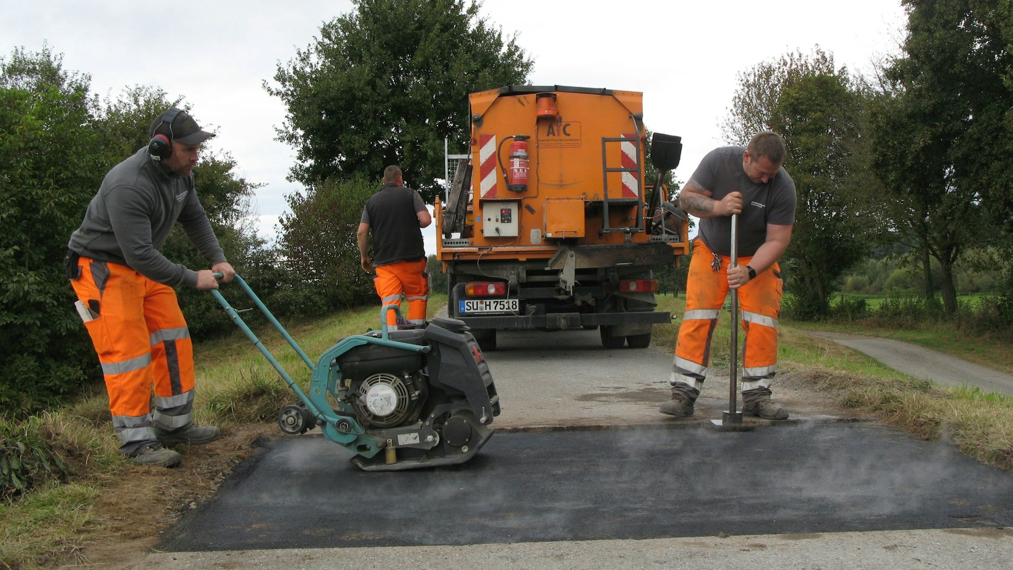 Drei Männer in orangefarbenen Arbeitshosen und grauen Oberteilen arbeiten mit Geräten und einem kleine Lastwagen auf dem Siegdeich.