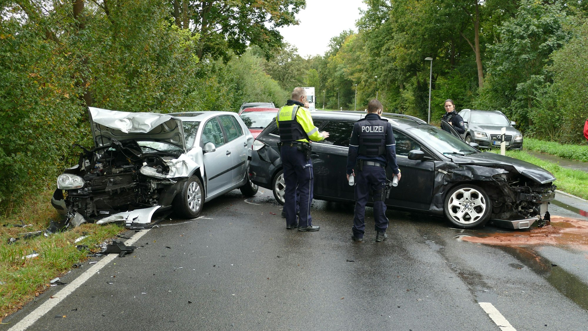 Ein silbernes und ein dunkles Auto stehen auf einer Straße, beide sind schwer beschädigt. Die Polizei steht neben den Fahrzeugen.
