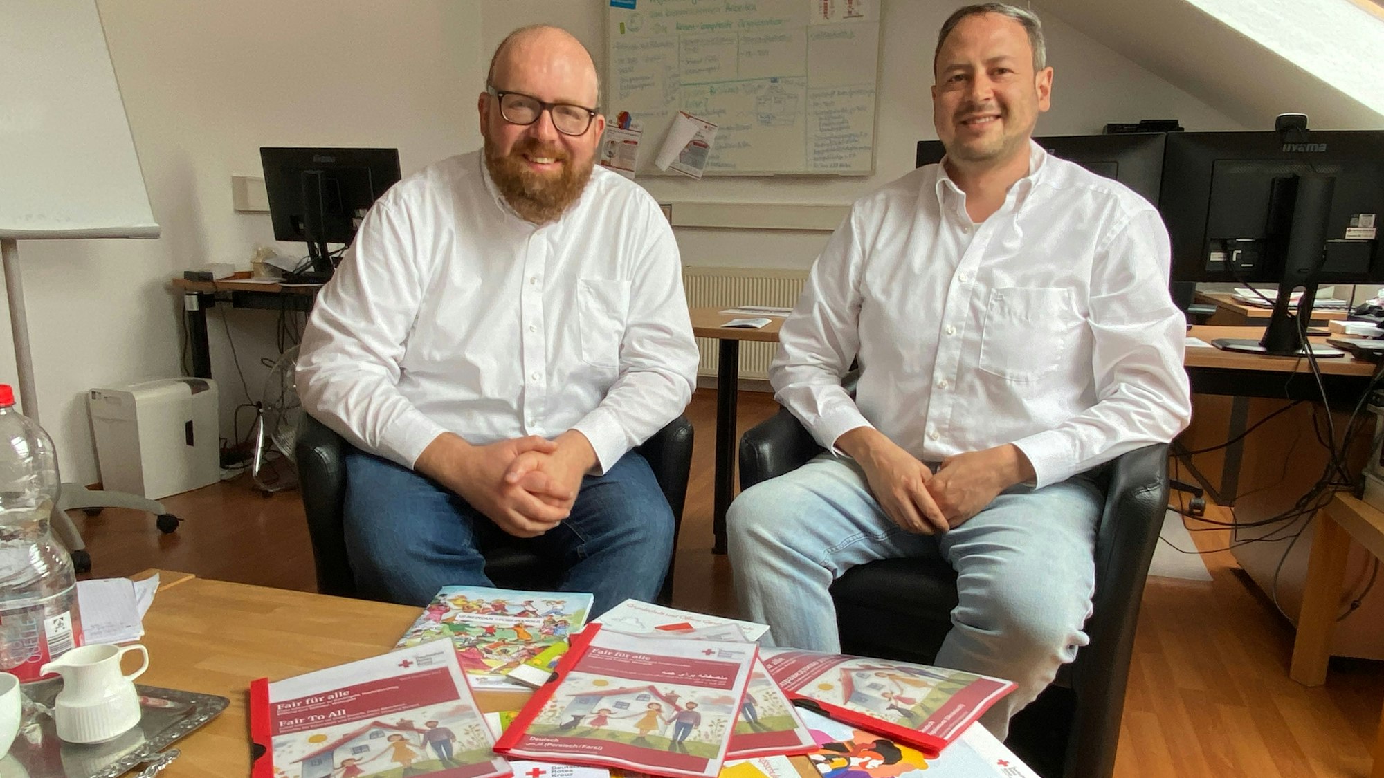 Zwei Männer sitzen vor einem Tisch, auf dem sämtliche mehrsprachige Broschüren der DRK-Integrationsagentur ausgelegt liegen.