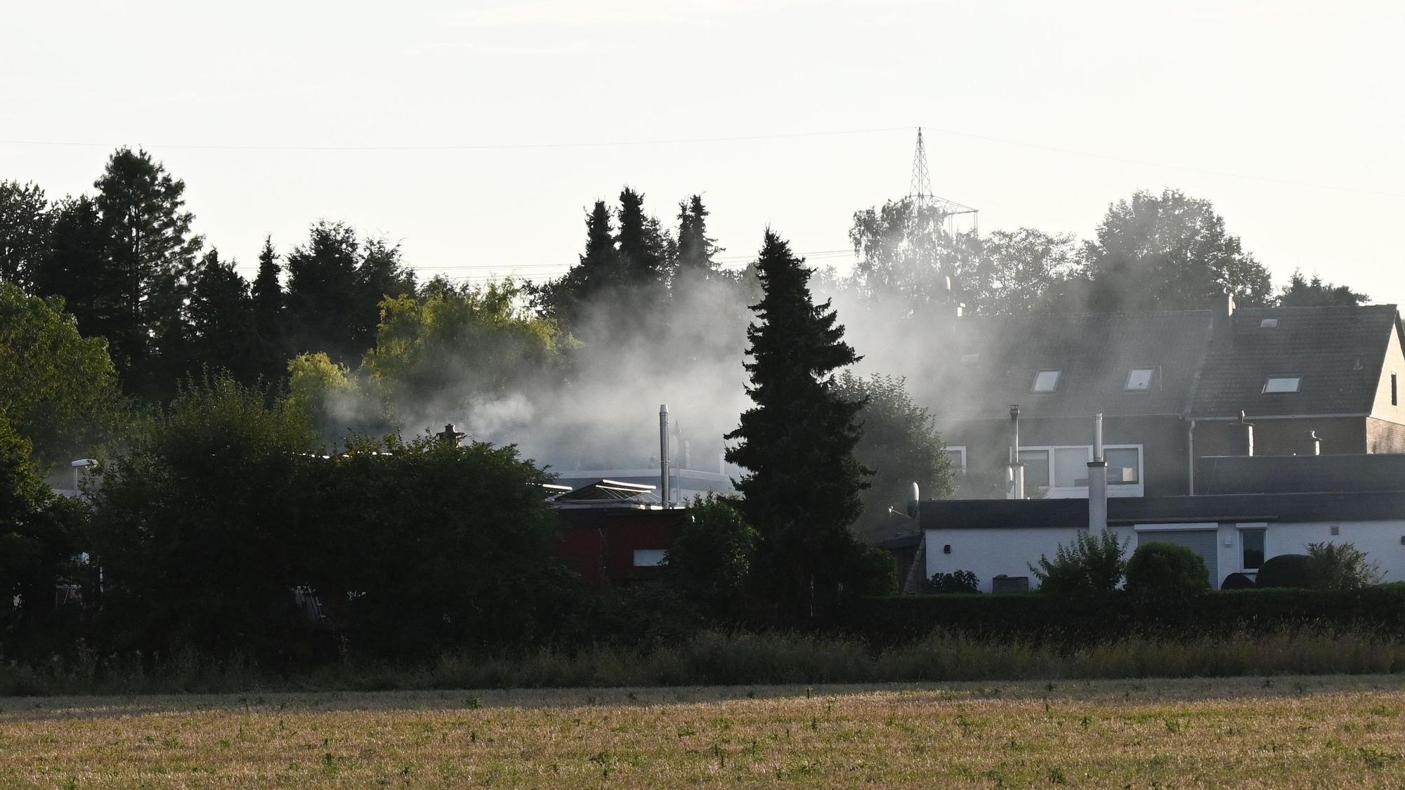 Ein Wohngebiet zwischen Büschen und Bäumen. Aus der Mitte steigt Rauch auf.