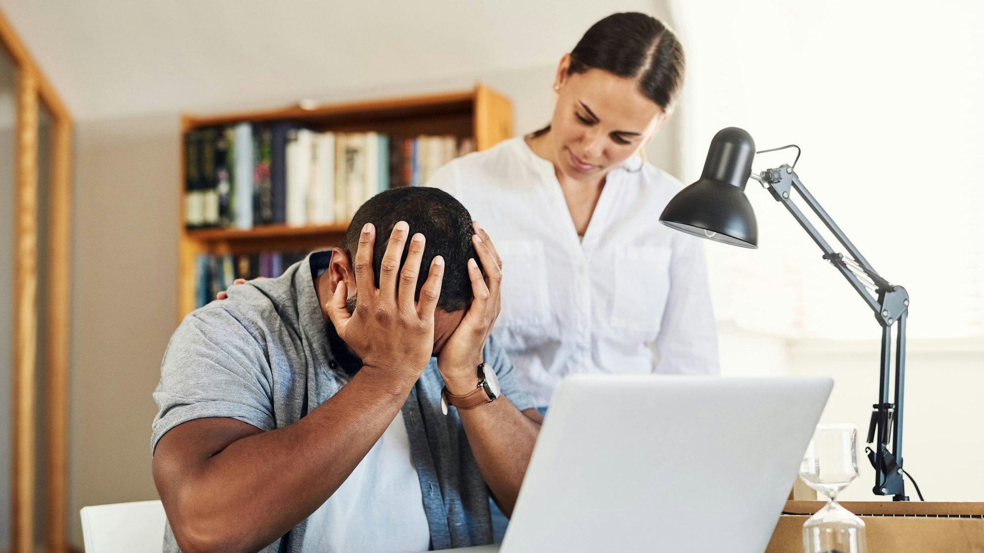 Mann sitzt am Schreibtisch und legt den Kopf in beide Hände, Frau steht daneben und legt ihre Hand auf seinen Rücken