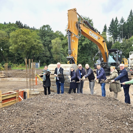 Vertreter der Unternehmen und aus Politik und Verwaltung mit Spaten in den Händen stehen vor einem Bagger.