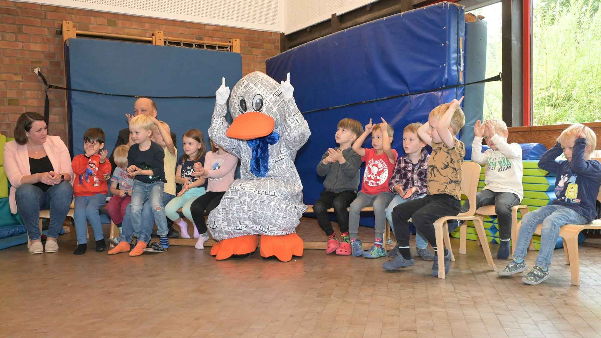 Kinder und Zeitungsente Paula Print sitzen auf Stühlen und einer Bank und singen.