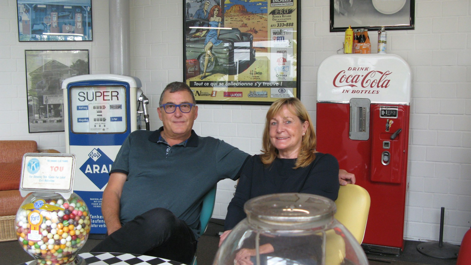 Ein Mann und eine Frau, Peter Weidenbrück und Gabi Stahl, sitzen auf Stühlen. An der Wand hinter ihnen stehen eine alte Tankstellen-Zapfsäule und ein alter Getränkeautomat.