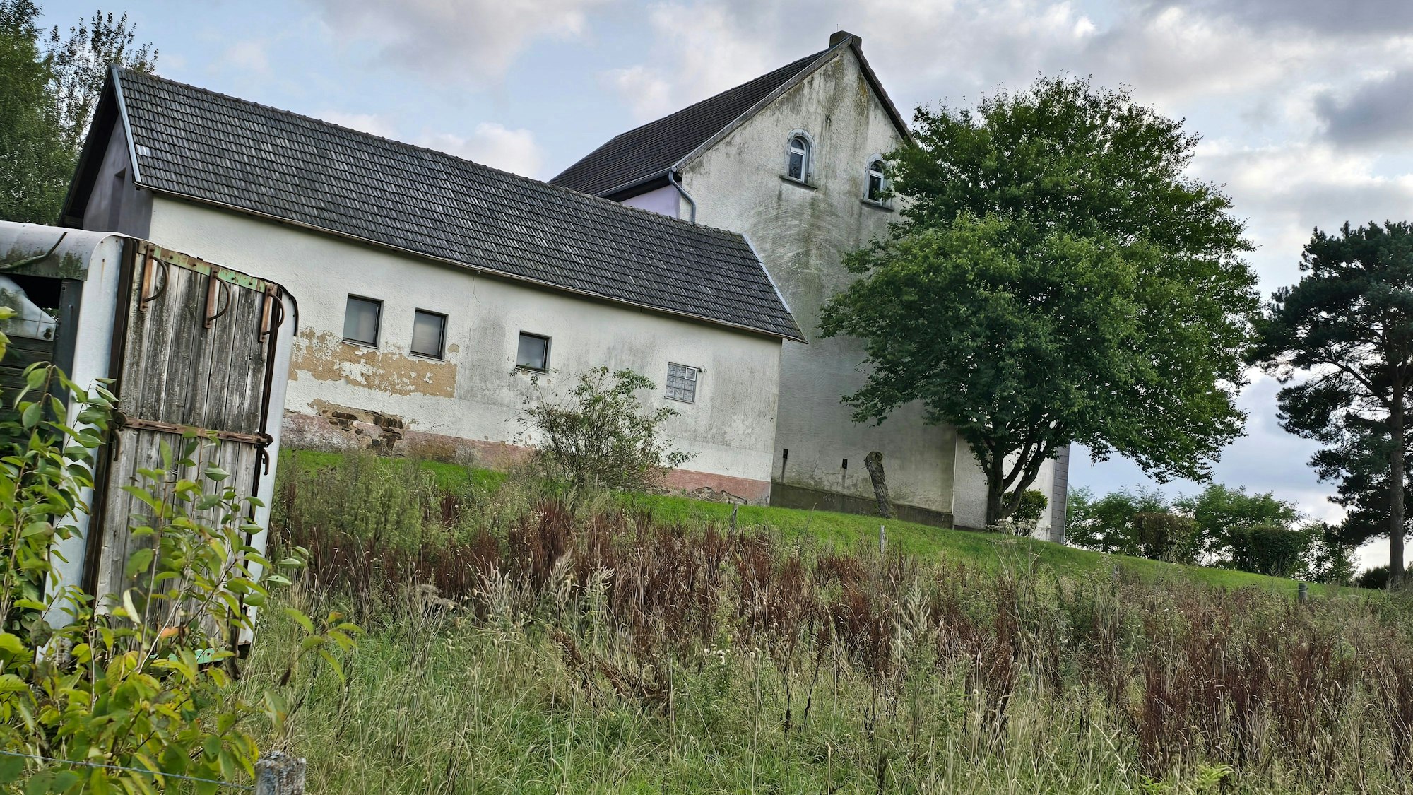 Ein etwas heruntergekommenes, weißes Haus steht auf einer Wiese, daneben Bäume.