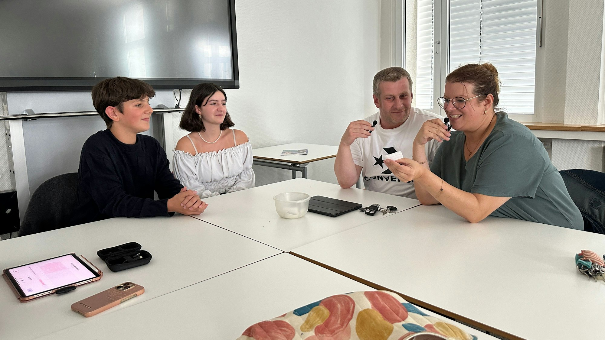 Zwei Schüler und zwei Lehrer sitzen gemeinsam an einem hellen Tisch, die Lehrerin rechts im Bild hat gerade ein Stück Papier aus einem Topf in der Mitte des Tischs gezogen.