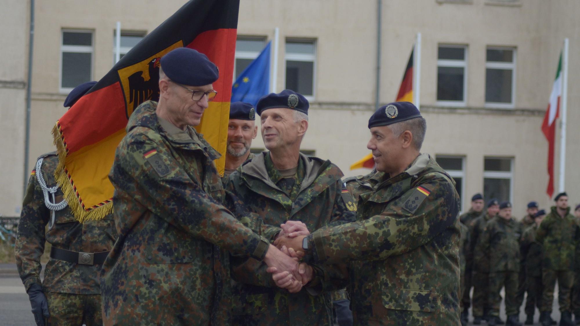 Die drei Offiziere reichen sich die Hände, hinter ihnen halten Soldaten die Truppenfahne.