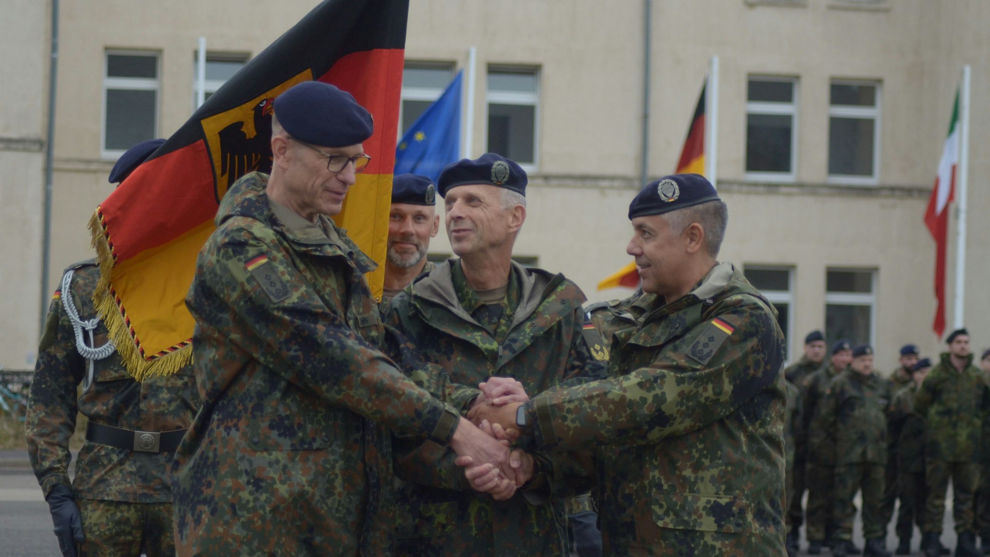 Die drei Offiziere reichen sich die Hände, hinter ihnen halten Soldaten die Truppenfahne.