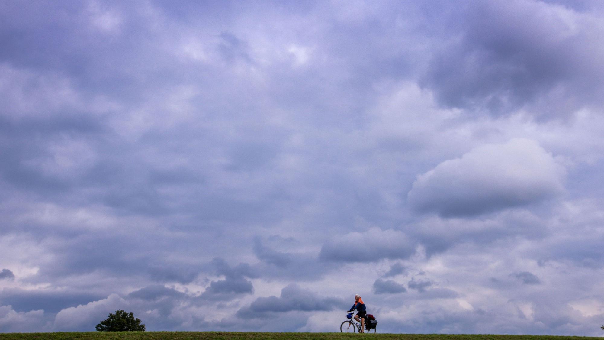 25.09.2024, Niedersachsen, Darchau: Unter einer dunklen Wolkenkulisse ist ein Radfahrer auf dem Elbdeich unterwegs. Mit sinkenden Temperaturen und dichter Bewölkung kündigt sich in Norddeutschland ein Wetterwechsel an. Foto: Jens Büttner/dpa +++ dpa-Bildfunk +++