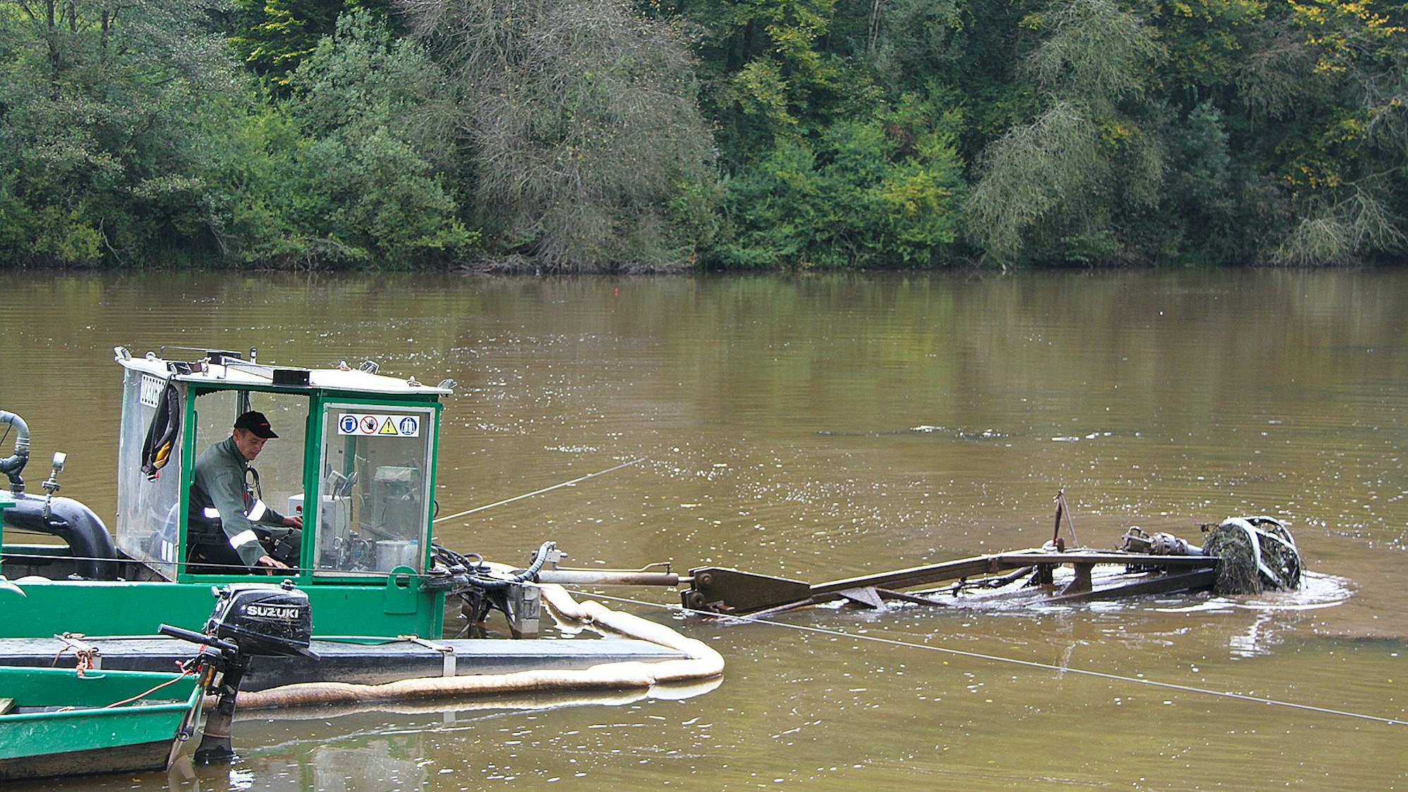 In einem Bagger mitten im Wasser sitzt ein Mann. Davor ist eine Vorrichtung angebracht, mit der der Schlamm abgetragen wird.