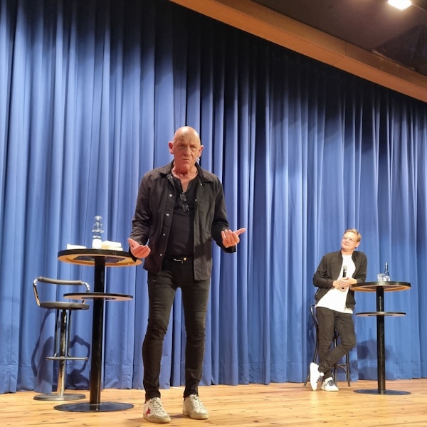 Der Schauspieler und Arzt Joe Bausch steht auf einer Bühne mit geöffneten Händen und gestikuliert in Richtung des Publikums.