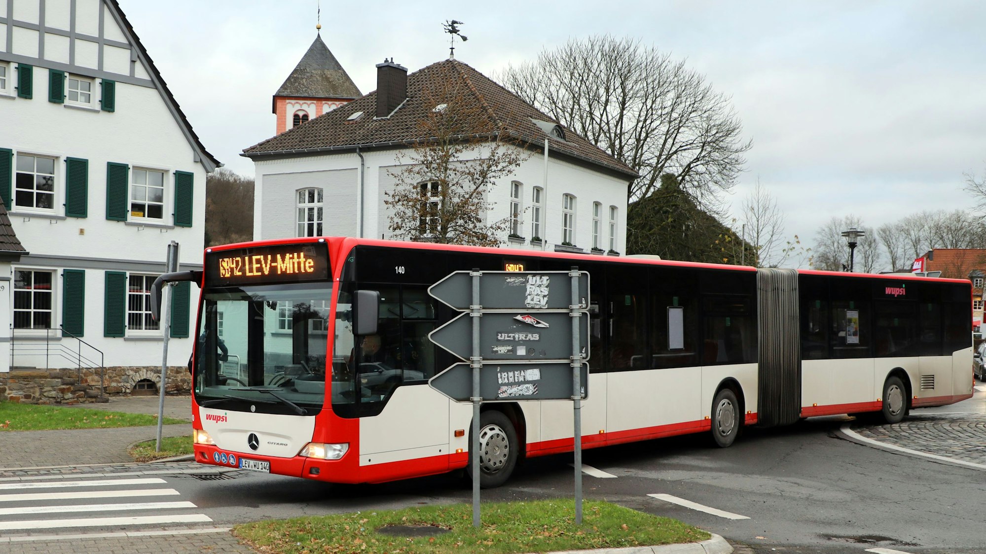 Ein Bus der Linie SB 42 fährt durch den Odenthaler Kreisverkehr. Im Hintergrund sind Rathaus und Kirche St. Pankratius zu sehen.
