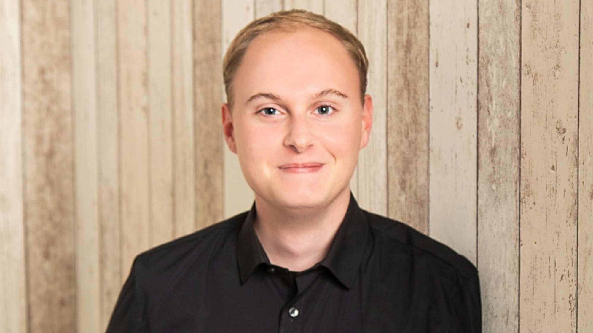 Ein Mann in schwarzem Hemd sitzt vor einer Wand aus Holz.