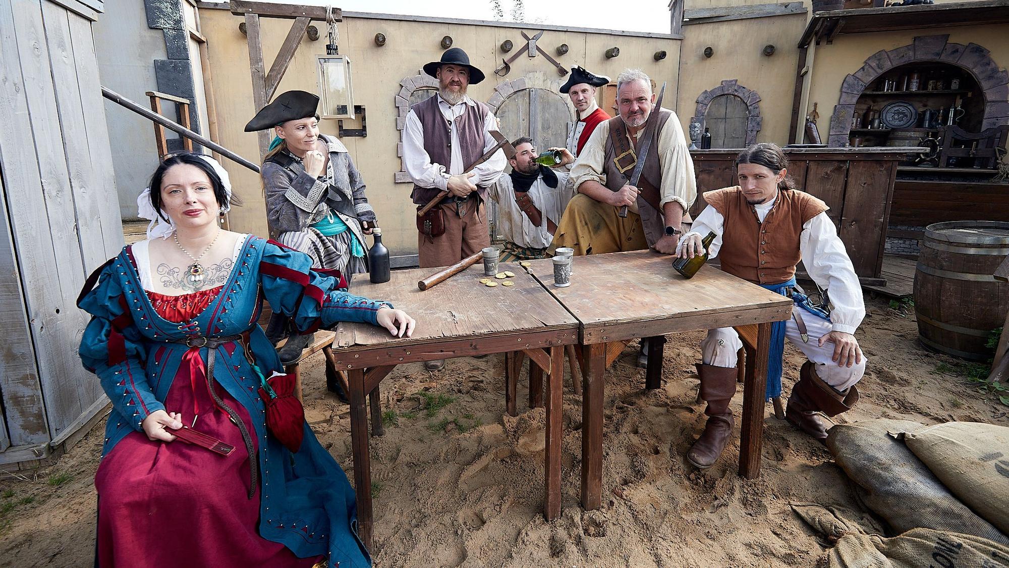 Menschen in Piratenkostümen sitzen an einem Holztisch, der auf sandigem Boden steht. Auf dem Tisch liegen Goldtaler, daneben stehen Trinkbecher.
