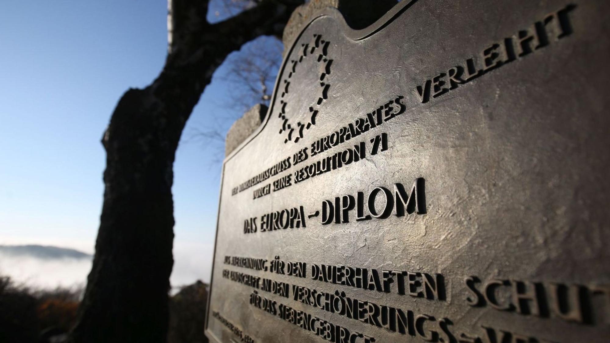 Eine Bronze-Platte mit der Aufschrift "Das Europa-Diplom", im Hintergrund ein Baum vor blauem Himmel.