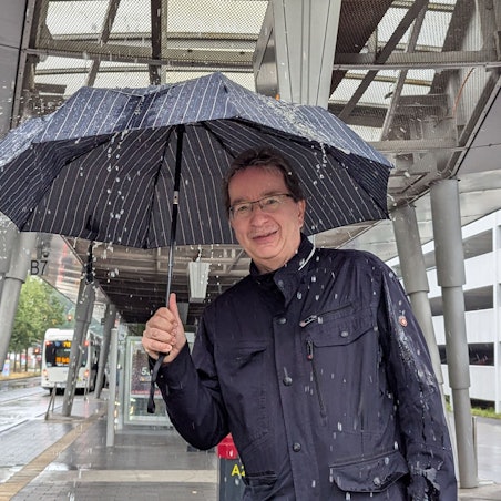 Das Foto zeigt einen Mann mit Regenschirm am Busbahnhof in Hürth Mitte. Auf den Schirm prasselt das Wasser.