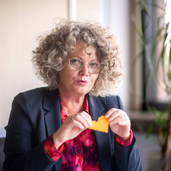 Eine Frau mit lockigem Haar und einer Brille im Anzug hält ein gebasteltes Herz in die Luft.
