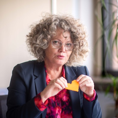 Eine Frau mit lockigem Haar und einer Brille im Anzug hält ein gebasteltes Herz in die Luft.