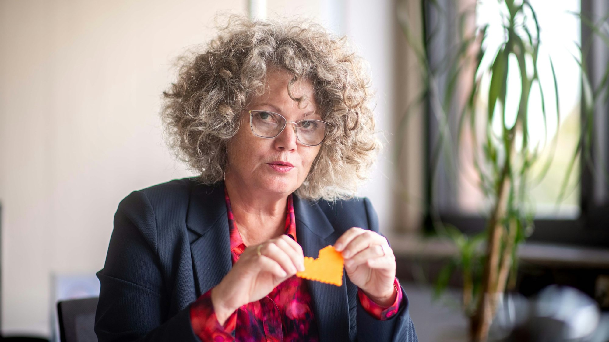 Eine Frau mit lockigem Haar und einer Brille im Anzug hält ein gebasteltes Herz in die Luft.